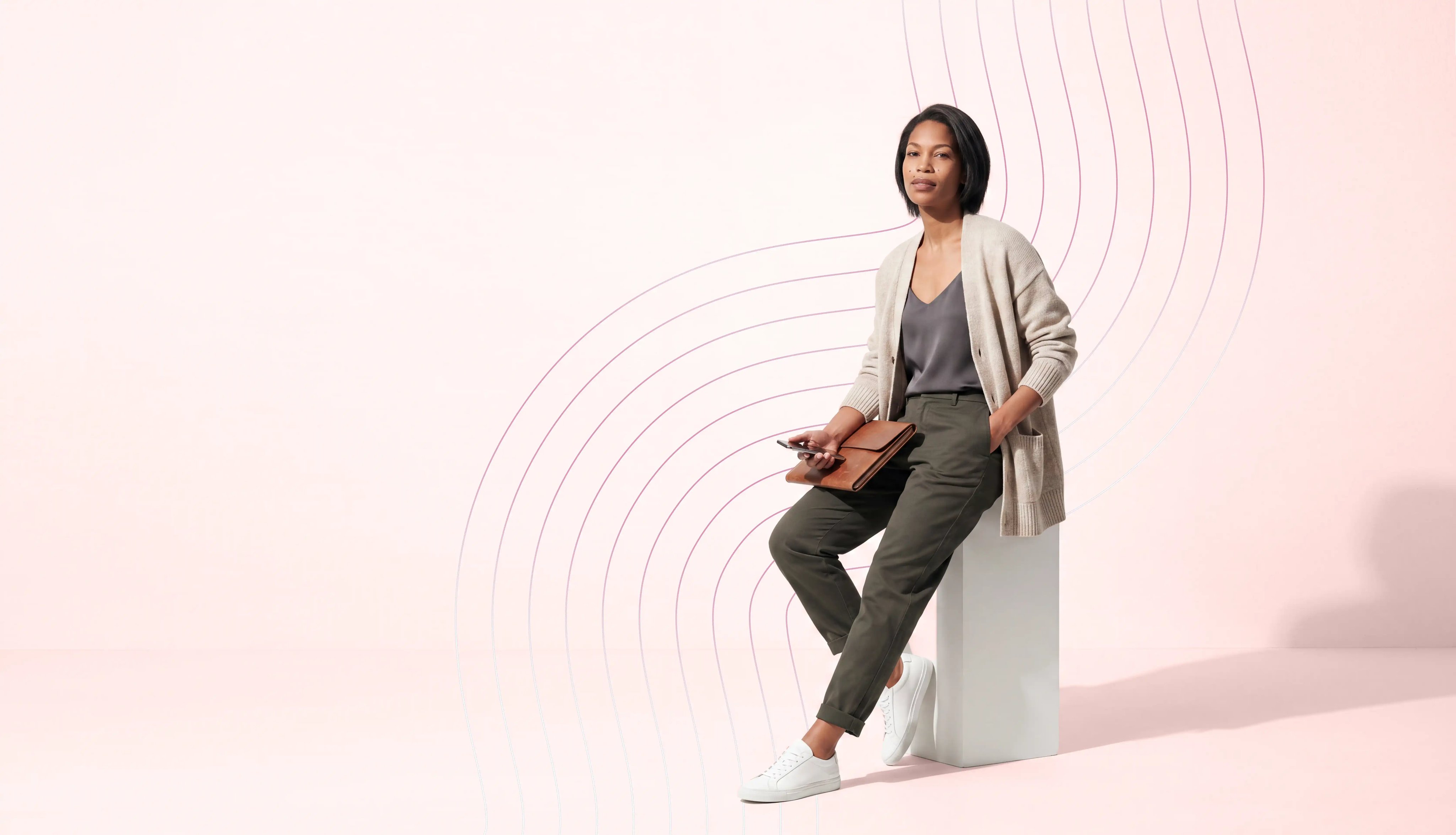 A woman wearing a cardigan sitting on a white stool on a pink background with geometric designs