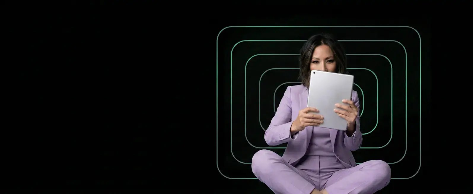 Woman wearing an all-purple suit sitting holding a tablet that covers most of her face against a black background with geometric designs