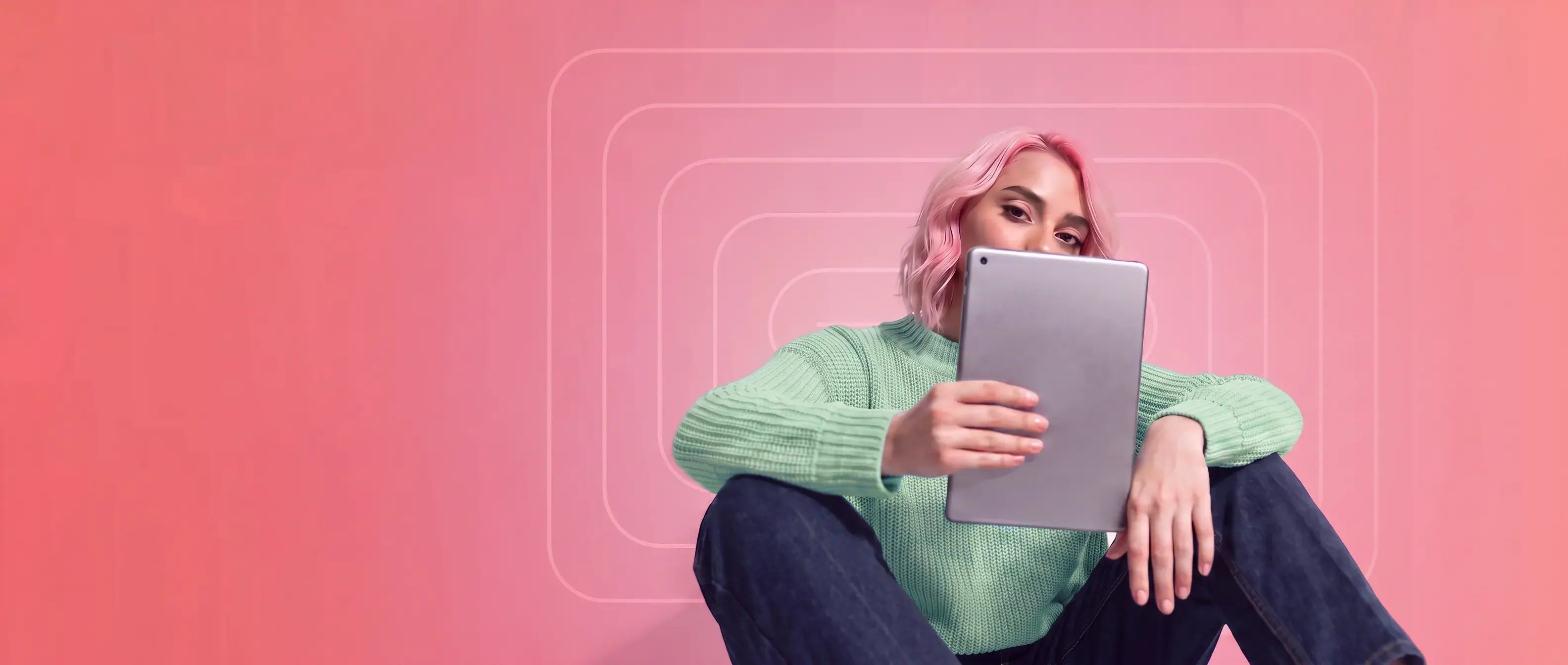 A woman wearing a green sweater holding a tablet over her face on a pink background with geometric designs
